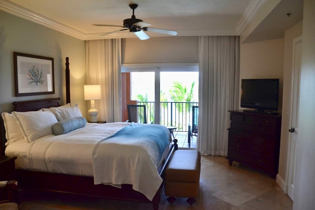 Une chambre d’hôtel équipée d’un lit, d’une télévision, d’une commode, d’un ventilateur de plafond et d’un balcon avec des rideaux.