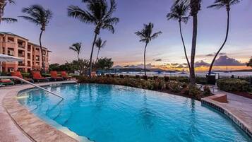 A pool with lounge chairs and palm trees, surrounded by a building with a view of the sunset.