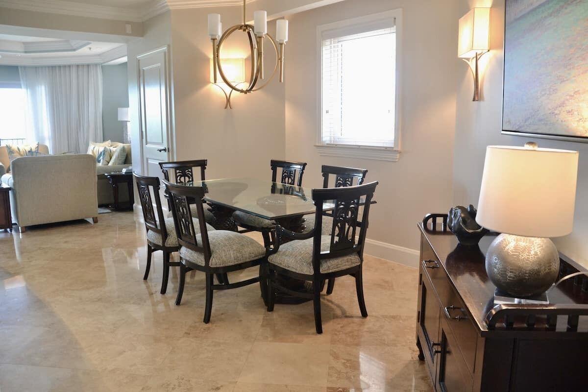 A dining area with a glass-top table and dark wooden chairs, a chandelier, and a lamp on a cabinet.