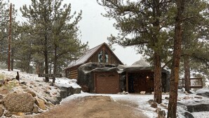 A rustic cabin with a dirt path leading to it, surrounded by snow-covered ground and pine trees.