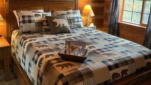 A wooden cabin bedroom with a bed featuring a quilt with bear patterns, a nightstand with a lamp, and a window with curtains.