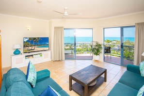 Relax in this stylish living room with a large flat-screen TV, two sofas, and a coffee table, offering a comfortable seating area and access to a balcony with ocean views.