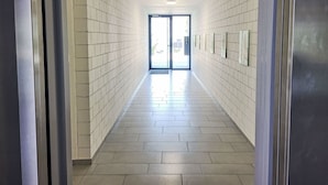 Un pasillo con paredes y piso de baldosas blancas, que conduce a una puerta de vidrio con vista al exterior.
