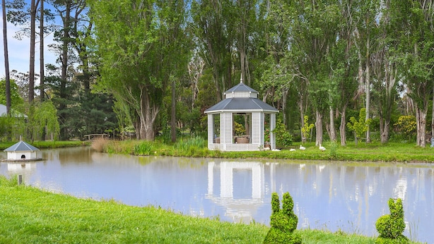 A gazebo by a pond with a small birdhouse and neatly trimmed bushes.