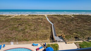 A beach view from a high vantage point, including a swimming pool with lounge chairs, a wooden boardwalk, and a sandy beach area. - Beachfront Bliss at Edgewater! Breath-taking Ocean Views, Pool & Hot Tub - Walk to Everything! (South Padre Island)