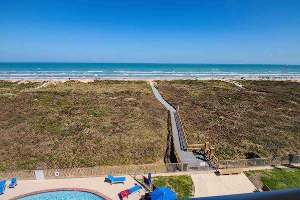 A beach view from a high vantage point, including a swimming pool with lounge chairs, a wooden boardwalk, and a sandy beach area.