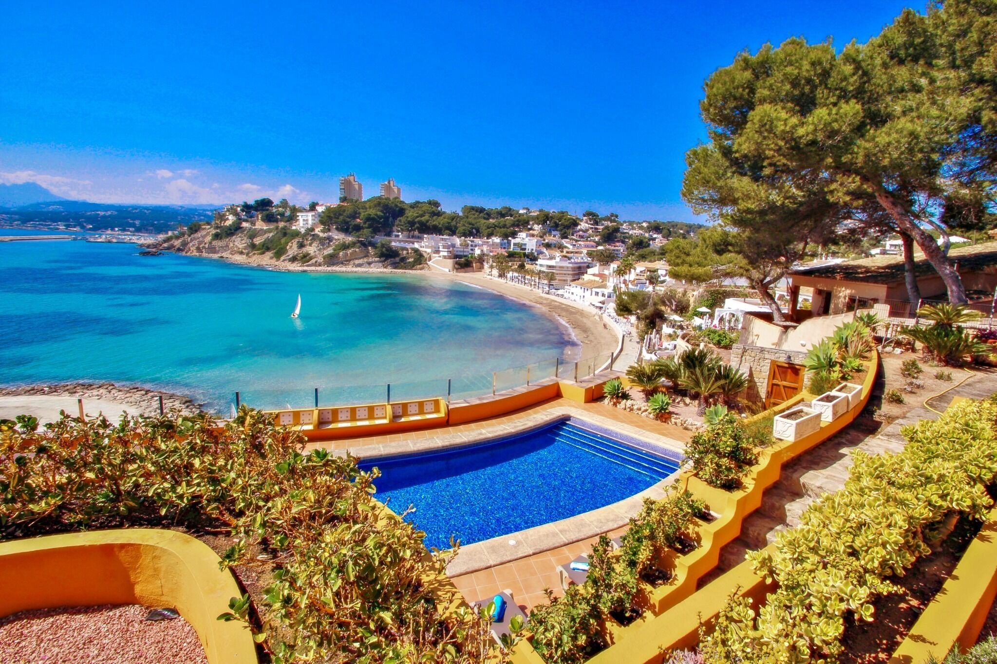 A coastal hotel with a swimming pool, beach access, and a view of the sea and distant buildings.