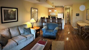 Una sala de estar con sofá, sillón y mesa de centro, un comedor con mesa y sillas, y una cocina con armarios y electrodomésticos.