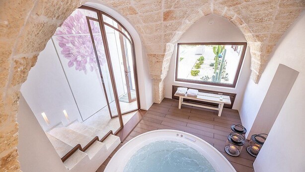 Un espace spa comprenant un bain à remous, des murs en pierre et un banc placé près d’une fenêtre donnant sur un jardin.