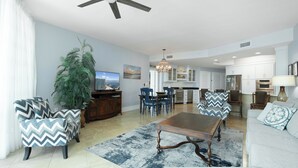 A spacious living area with a ceiling fan, a large potted plant, a television, and a dining area with chairs and a table. - Two Bedroom Condo with Views of Gulf, Bay, and Outdoor Pool Decks! (Orange Beach)