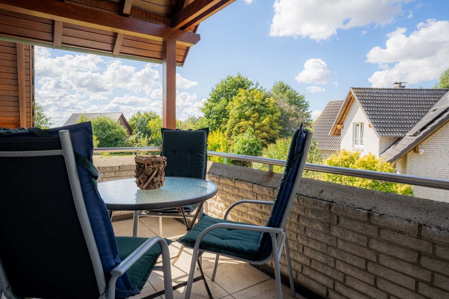 Ein überdachter Außenbereich mit einem runden Tisch, zwei Stühlen, einem dekorativen Holzstück auf dem Tisch und Blick auf Häuser und Bäume im Hintergrund.
