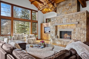 A spacious living room with a stone fireplace, leather sofas, and large windows offering a view of trees.