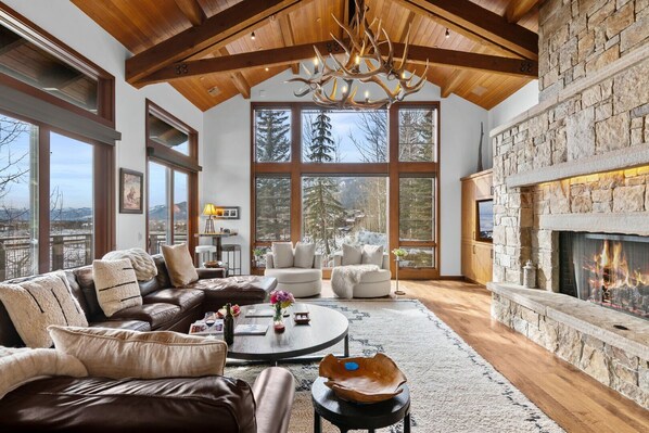 A spacious living room with a fireplace, large windows, and a high ceiling with wooden beams.