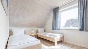 Quarto com teto inclinado, duas camas, mesa de cabeceira e janela com cortinas.
