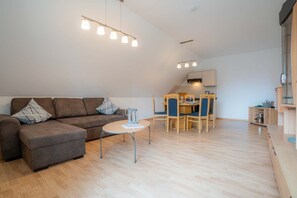 A modern living room with a brown sofa, a small round table, and a dining area with wooden chairs and a kitchen in the background.