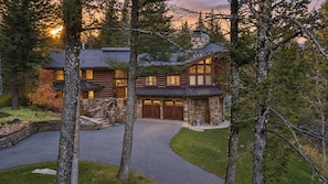 A log cabin with a stone exterior, a stone chimney, and a driveway leading to a gated entrance.
