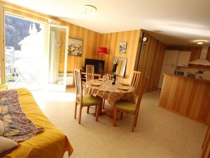 A cozy room with a dining table set for two, wooden walls, a small kitchen area, and a bed with a yellow cover.
