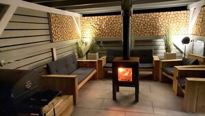 Un espace de détente extérieur accueillant, doté d’un poêle à bois, de sièges confortables et d’un mur en pierre en arrière-plan.