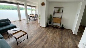 Un vaste espace de vie doté d’un plancher en bois, d’un canapé, d’une table basse et d’une table à manger avec des chaises. Une grande fenêtre offre une vue sur un lac et la verdure environnante. On peut également apercevoir une horloge et une étagère à plantes.