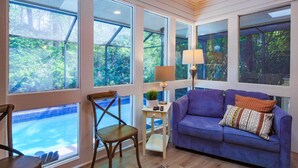 Une pièce dotée de grandes fenêtres, d’un canapé violet, d’une chaise en bois et d’une petite table sur laquelle se trouvent une lampe et une plante.