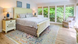 Une chambre à coucher comprenant un lit, des tables de chevet, une commode, une fenêtre donnant sur un espace verdoyant et un fauteuil.