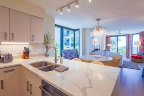 Una cocina moderna con encimera de mármol, pileta de acero inoxidable y lavavajillas integrado. Hay un comedor con una mesa redonda y sillas, y una sala de estar con sofá y un tapete colorido.
