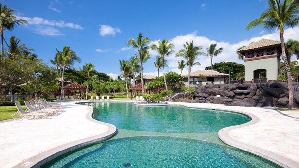 A clear swimming pool with lounge chairs, palm trees, and a building with a tiled roof.