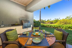 An outdoor kitchen with a grill, dining table set with green plates, bowls, and utensils, surrounded by wicker chairs with green cushions.