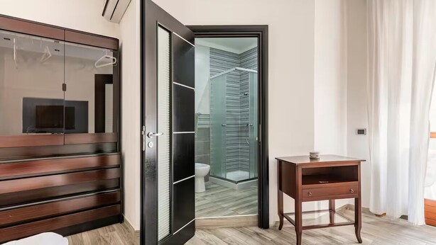 Una habitación de hotel moderna con un armario de madera, una puerta de vidrio que da al baño y una mesita de noche de madera.