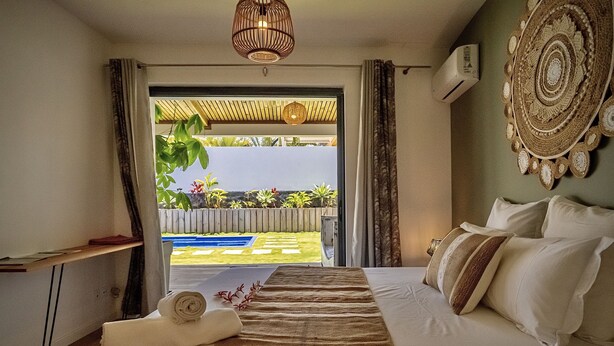 Ein Hotelzimmer mit einem Bett, einem Nachttisch, einem Handtuch, einer Hängelampe und Blick auf einen Pool und Grünflächen.