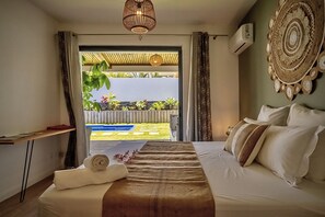 Une chambre d’hôtel avec un lit, une table de chevet, une serviette, un luminaire suspendu et une vue sur une piscine et la verdure environnante.