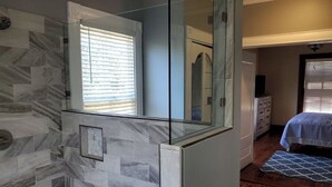 Ein Badezimmer mit Duschkabine aus Glas, einer Wand mit Marmoroptik und im angrenzenden Raum ein Bett mit blauer Bettdecke.