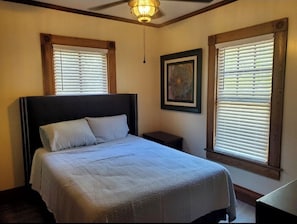 Un dormitorio con una cama grande, dos mesitas de noche, un ventilador de techo y ventanas con persianas.