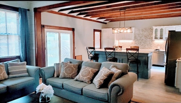 Una sala de estar con un sofá, una mesa de centro con flores, y una zona de cocina con una mesa de comedor y sillas.
