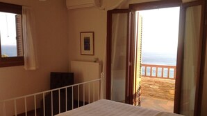 Ein Schlafzimmer mit Bett, einem Fenster mit Jalousien, einem Heizkörper und einer Tür, die auf einen Balkon mit Meerblick führt.