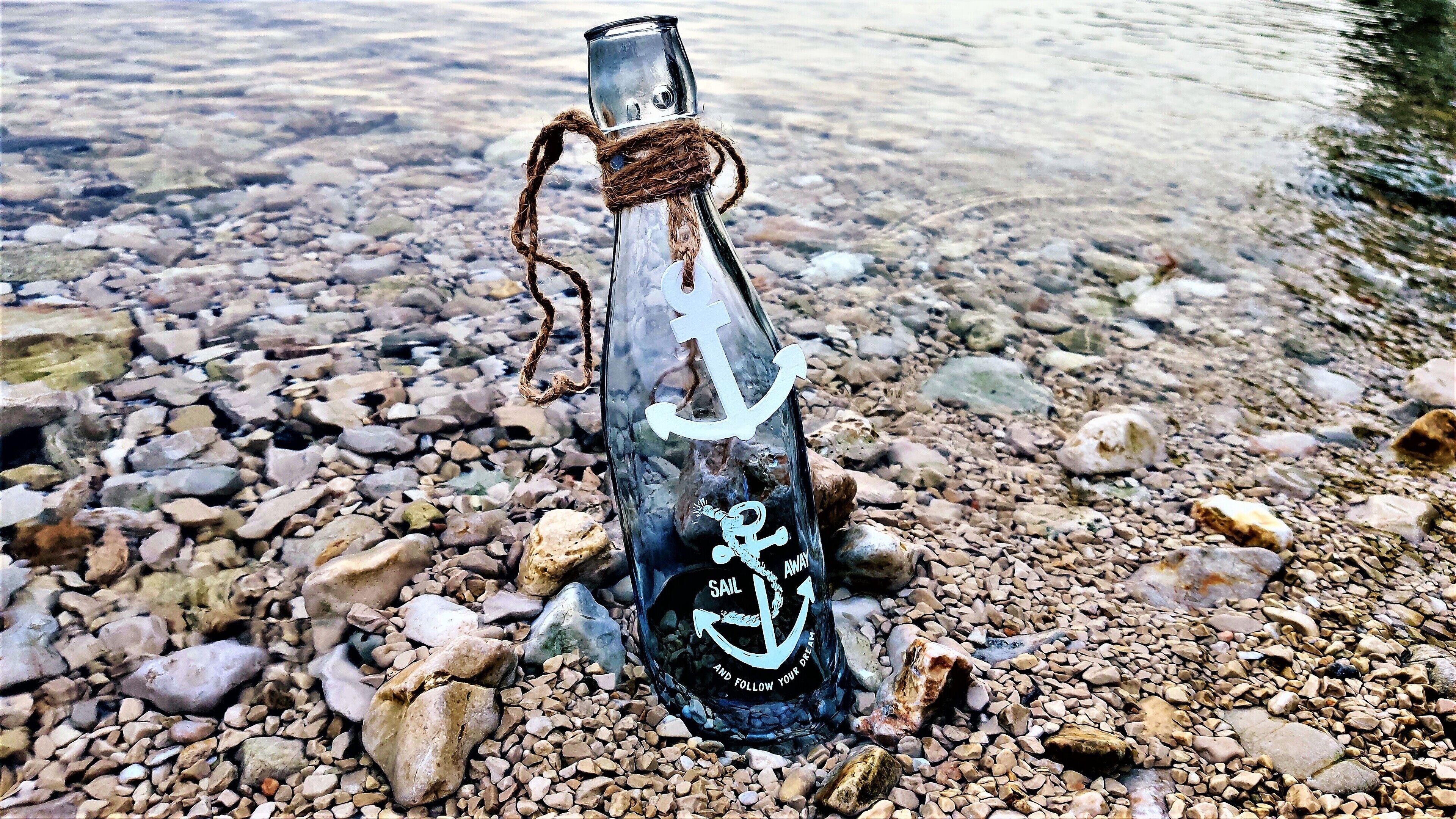 A clear glass bottle with a rope tied around its neck, placed on a rocky shore with clear water in the background.