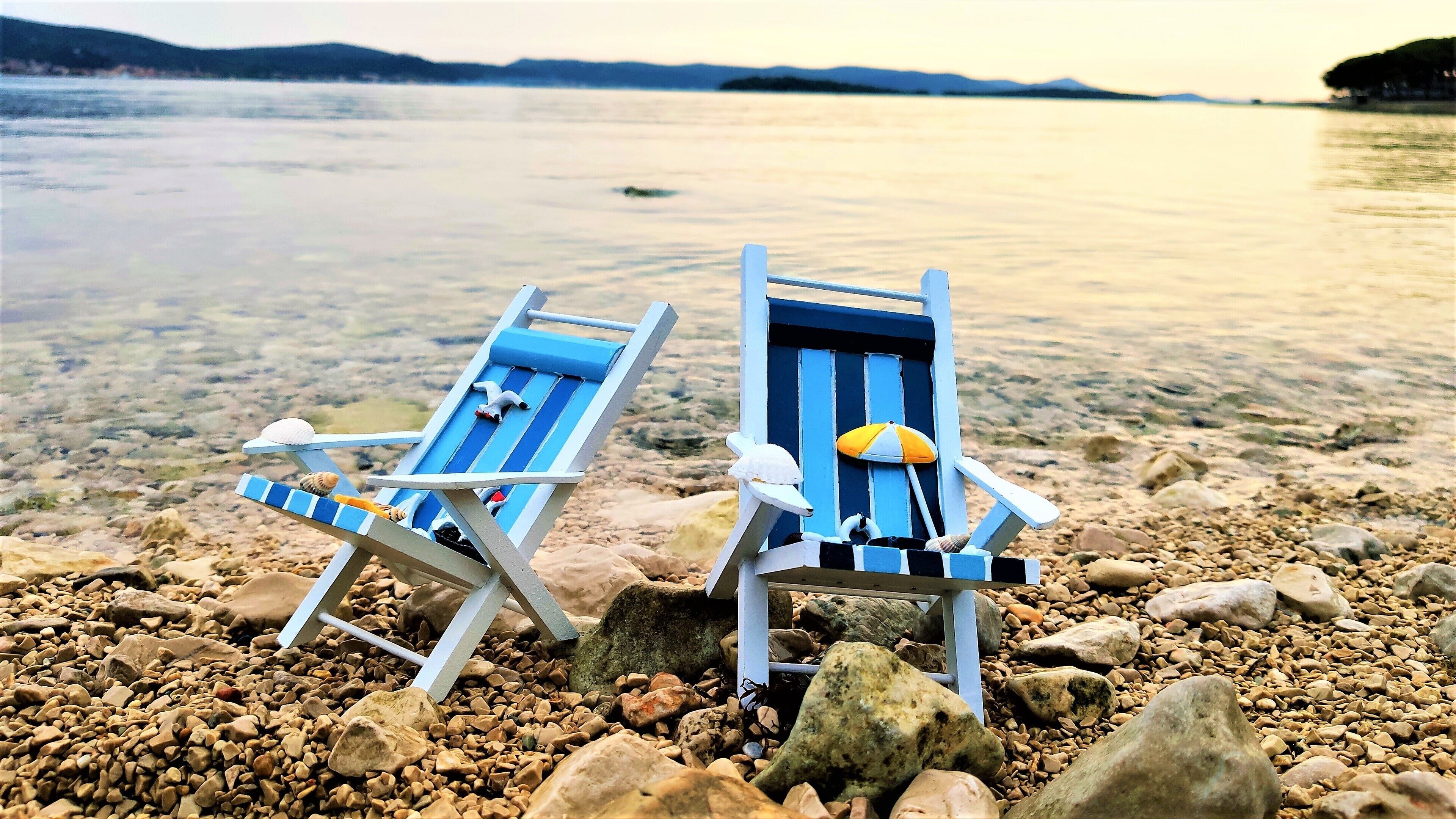Deux chaises de plage sur une plage de galets, avec un lac paisible et des collines lointaines en arrière-plan.