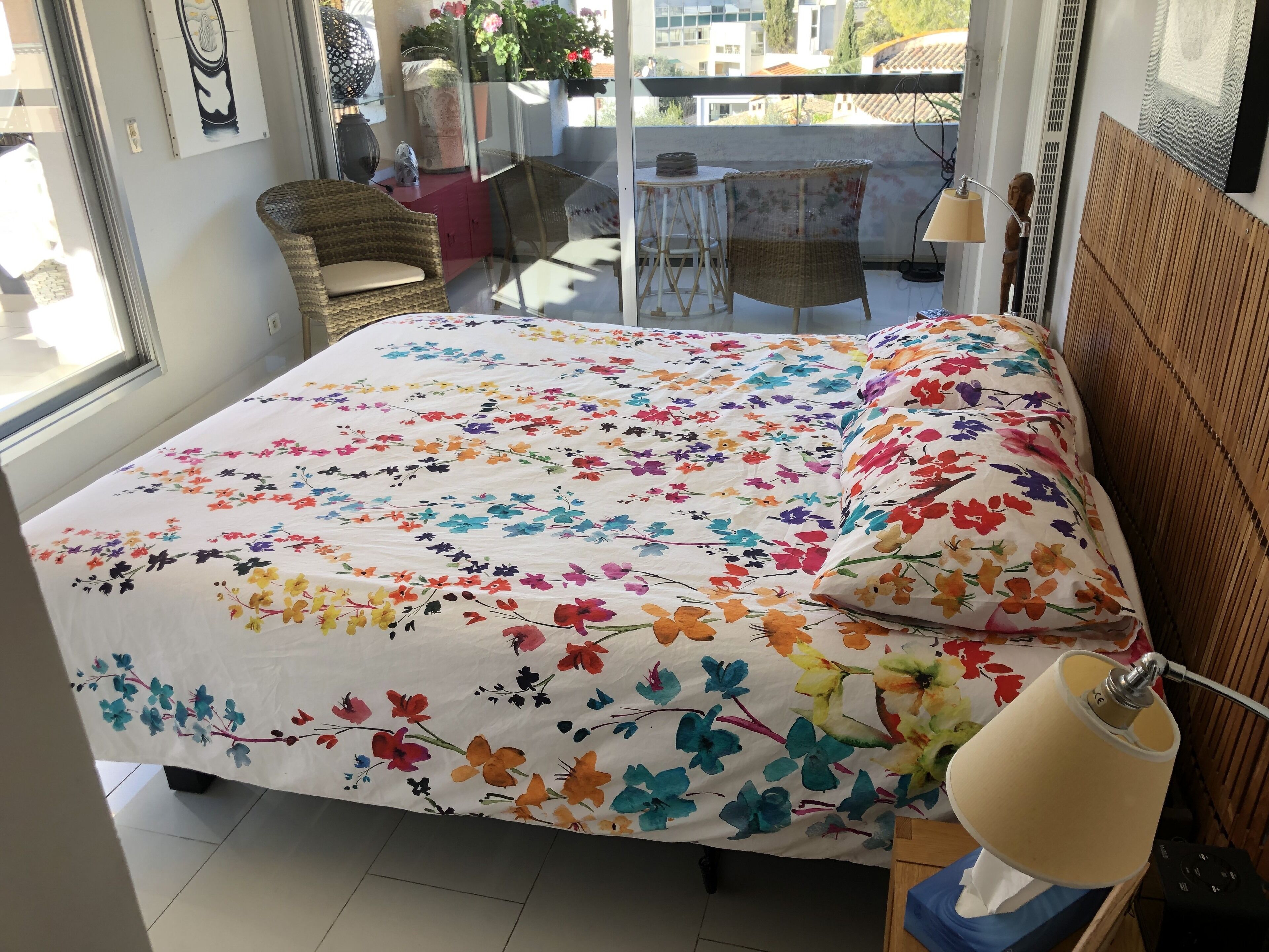 Une chambre avec une couvre-lit à motifs floraux, une tête de lit en bois, une lampe de chevet et une vue sur le balcon, qui est aménagé avec du mobilier d’extérieur.