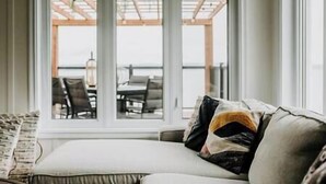 A room with a sofa facing a large window overlooking a terrace with outdoor furniture. - Oakes House+waterfront+games room+hot tub+Firepit (Charlo)