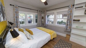 Quarto com cama, luminária de cabeceira, ventilador de teto, janelas, cortinas e uma estante de livros.