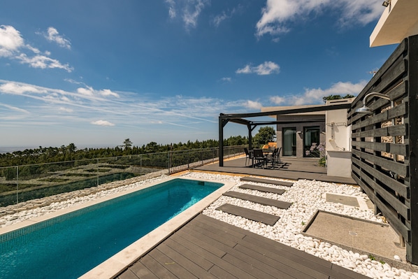 A swimming pool with a wooden deck, surrounded by white pebbles, and a covered seating area with a view of the landscape.