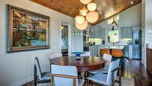 Uma área de refeições com mesa e cadeiras de madeira, um quadro grande na parede e uma cozinha ao fundo.