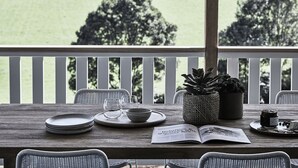 A wooden table with plates, glasses, a bowl, a book, and a potted plant on a balcony with a view of a green field and trees. - Benny’s Cottage  Luxury hinterland accomodation & photo shoot location (Clunes)