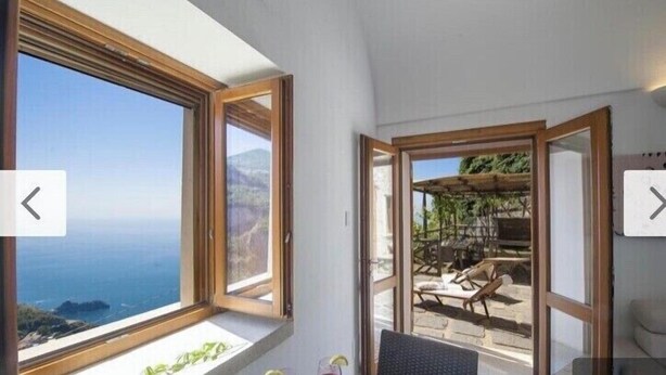 Une pièce dotée d’une grande fenêtre donnant sur la côte, et d’une porte-fenêtre coulissante qui mène à un espace extérieur aménagé avec une pergola.