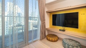 Een moderne woonkamer met een groot raam, een flatscreen televisie bevestigd aan een gele muur en een gestreept voetenbankje.