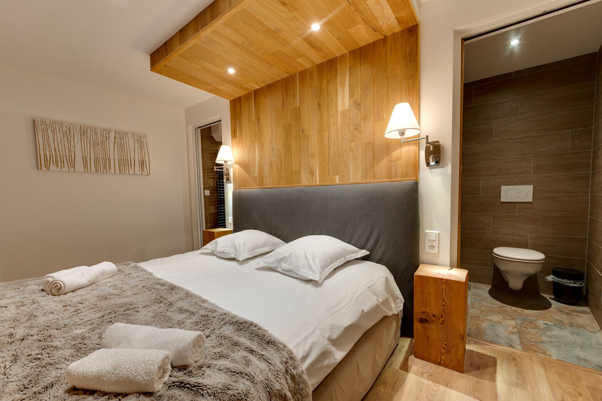 Een moderne hotelkamer met een groot bed, een houten hoofdeinde, een wandlamp en een zichtbare badkamer.