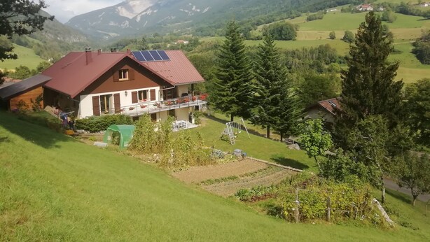 A house with a red roof and solar panels, surrounded by green fields and mountains.