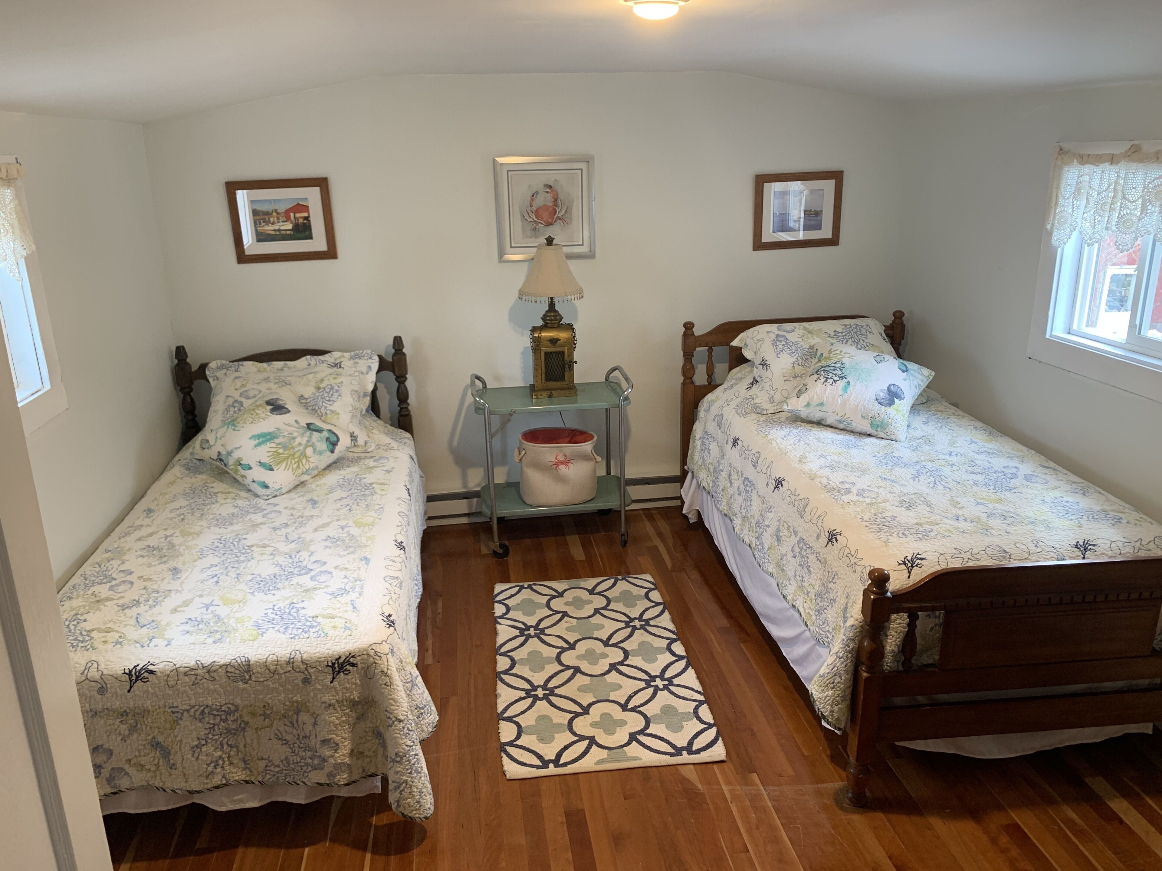 A bedroom with two single beds, a nightstand with a lamp, a small table with a pot, and a patterned rug.