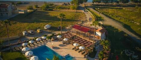 An aerial view of a resort with a swimming pool, lounge areas, and a view of the sea.