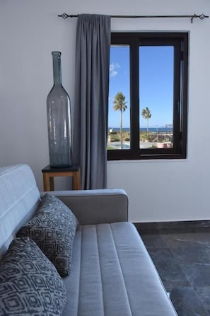 A room with a sofa, a large window, and a view of palm trees and a building. - Odos Oneiron Tavronitis Bay (Platanias)